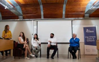 Imagen de expertos que expusieron en el curso de epilepsia, dentro de la sala 705 de la Facultad de Administración y Economía. A la izquierda, una mujer de pie con blusa amarilla, al lado de ella una mujer sentada con blusa sin mangas blanca y falda negra. Al centro está una mujer de blusa blanca manga larga y pantalón con rayas blancas y negras. Le sigue un hombre con barba y lentes, vestido con camisa blanca y pantalón negro. En el costado derecho un hombre canoso, con lentes, vestido con chaqueta azul