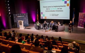 Imagen del escenario de Auditorio FAE con cuatro mujeres sentadas en unos sillones al centro. Hay una pantalla de fondo con un lienzo blanco y el texto en negro que dice Claves para la implementación de la Ley Karin en las Universidades Estatales.