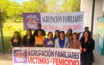 Grupo de personas ubicadas afuera de casa central, principalmente mujeres incluida la Vicerrectora de Calidad de Vida, Género, Equidad y Diversidad, doctora Karina Arias quien está al centro., que portan un lienzo que dice Agrupación Familiares de Víctimas de Femicidio