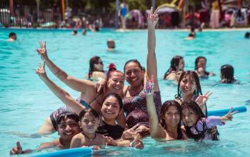 Grupo de personas, hombres, mujeres, niños y niñas, al borde de la piscina y levantando las manos en señal de alegría.