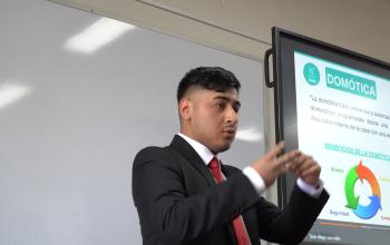 Estudiante vestido formal, con chaqueta oscura y corbata roja expone ante la comusión con una pantalla de fondo que muestra su tesis