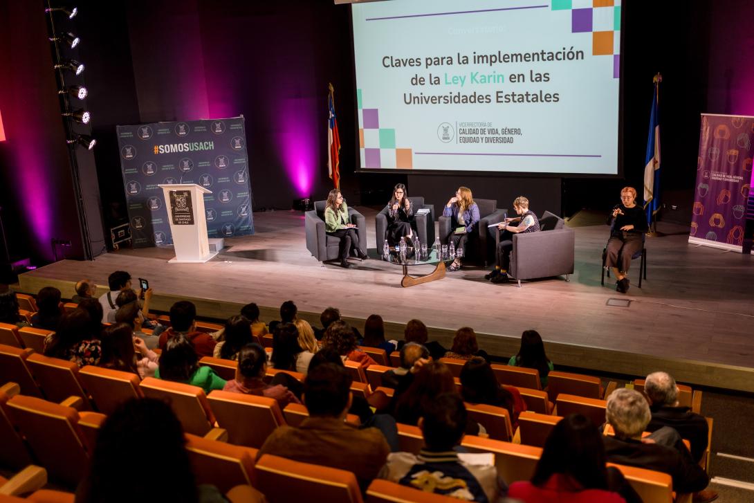 Imagen del escenario de Auditorio FAE con cuatro mujeres sentadas en unos sillones al centro. Hay una pantalla de fondo con un lienzo blanco y el texto en negro que dice Claves para la implementación de la Ley Karin en las Universidades Estatales.