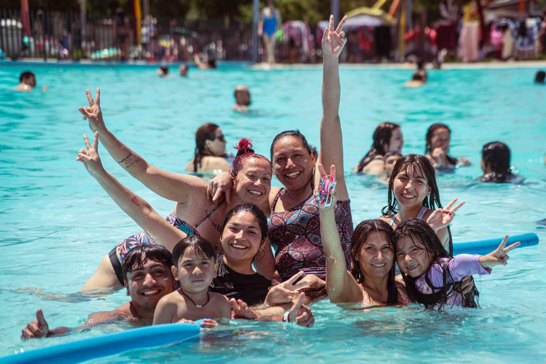 Grupo de personas, hombres, mujeres, niños y niñas, al borde de la piscina y levantando las manos en señal de alegría.